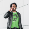 A happy looking human holds a phone while wearing a green shirt with a picture of Louis Riel and the words "Keepin' It Riel"