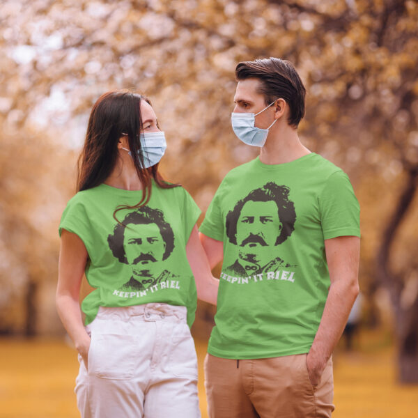 2 humans wear covid masks while also wearing shirts with a picture of Louis Riel and the words "Keepin' It Riel"